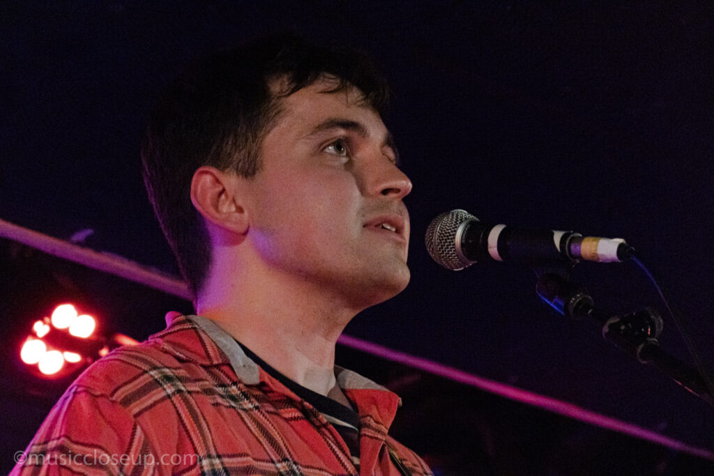 A close-up photo of Callum Pitt in front of a microphone. He is wearing a red checked shirt.