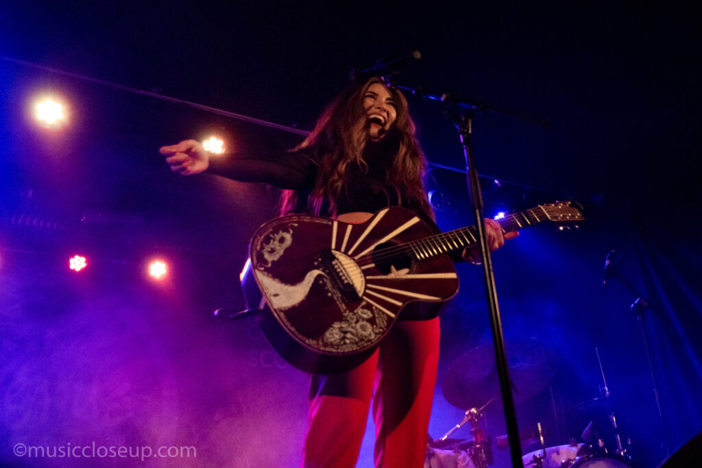 Tenille Townes live in Glasgow: Tenille laughs as she plays guitar. the pattern of the guitar is visible. It is a white sunburt pattern with a path and flowers. 