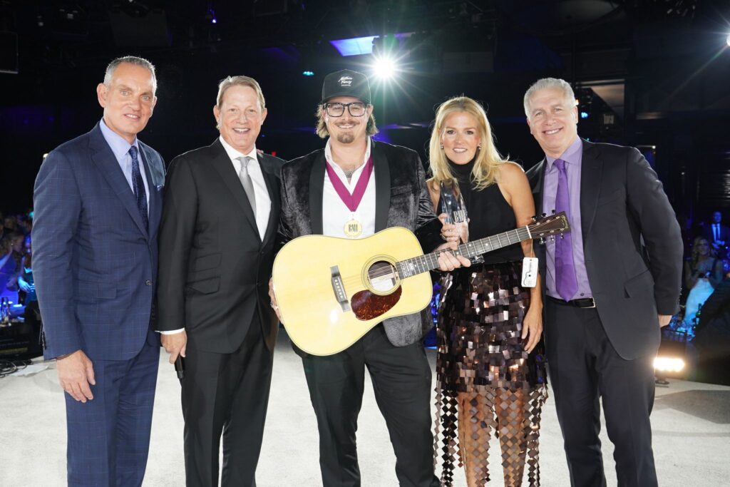 HARDY wearing a medal, a baseball cap and an acoustic guitar. He is standing with three men and a woman at the BMI Country Awards 2022.