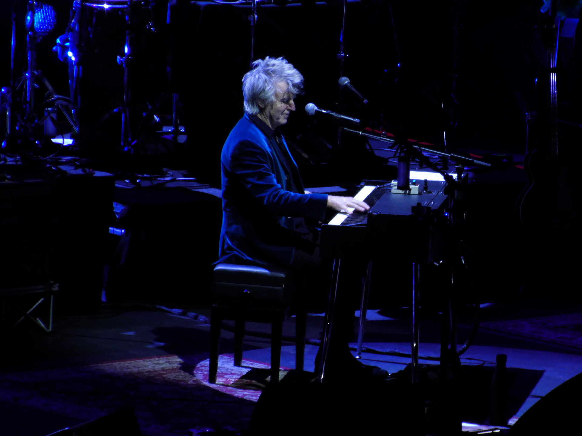 Neil Finn, dressed in a navy velvet jacket, playing a piano with Crowded House at Glasgow Hydro