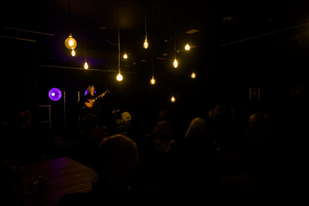 Emily Barker playing guitar in a darkened room with lightbulbs hanging from the ceiling