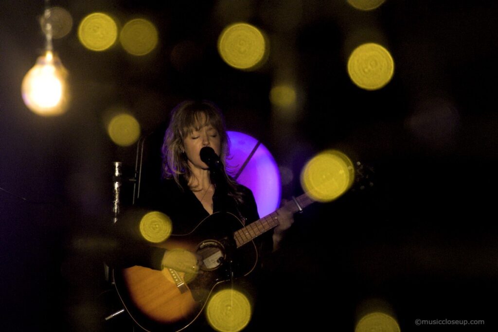 Emily Barker performing a soke surrounded by a bokeh effect from hanging lightbulbs