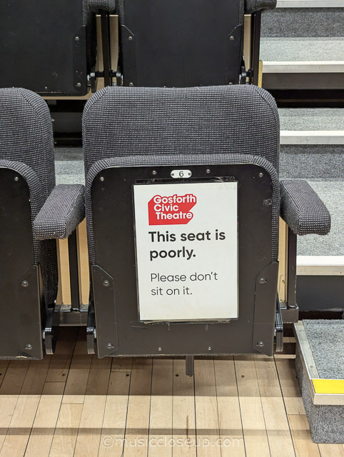 A grey chair with a sign that reads 'This seat is poorly. Please don't sit on it.'